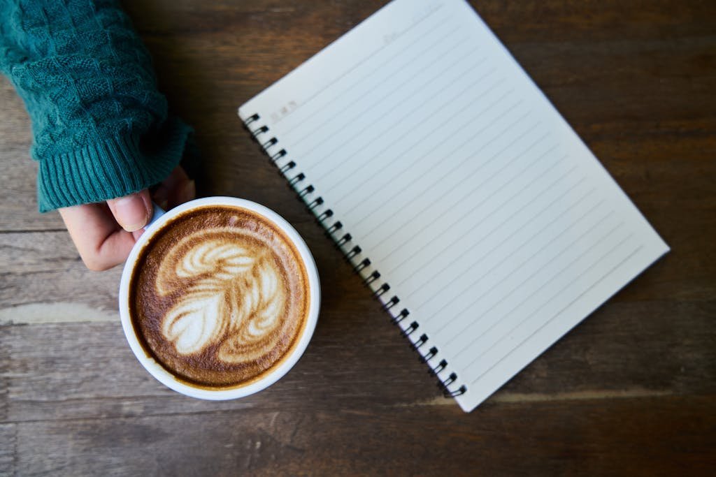 Top view of a latte with latte art next to a blank notebook on a wooden table.